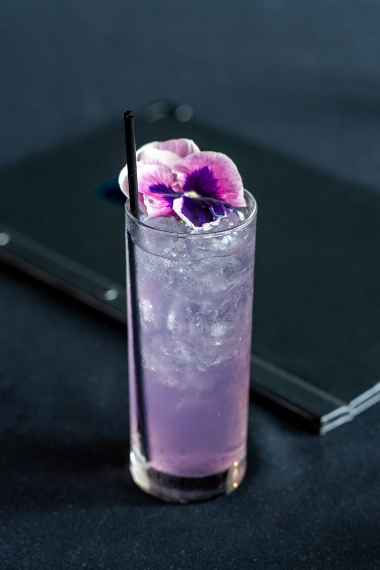 An award-winning vibrant purple cocktail, topped with a purple hibiscus flower. Served in a glass with a black stirring straw.
